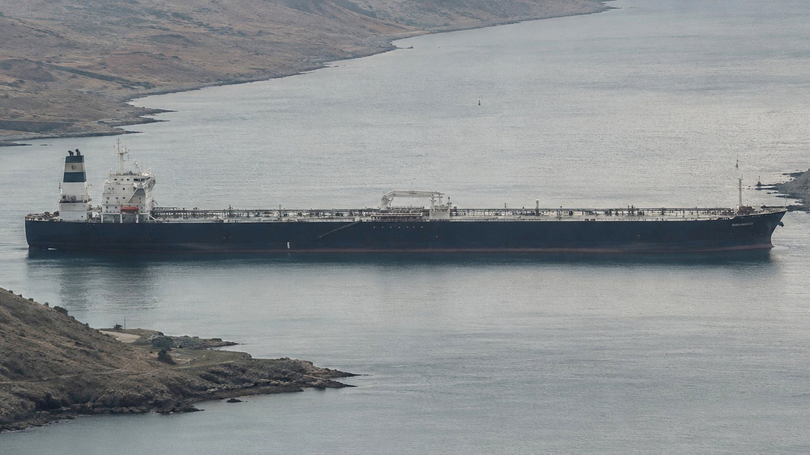 A lone oil tanker sitting still in the grey, overcast water of a narrow strait, the arid chokepoint coastline pressing in on both sides.