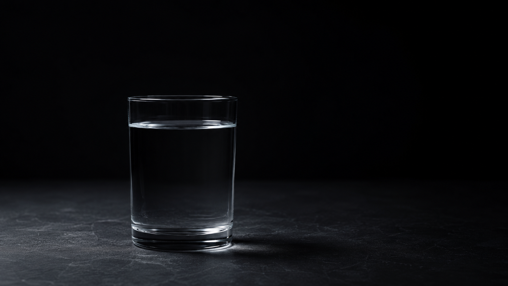 A plain glass of clear water on a dark surface, lit low from one side.